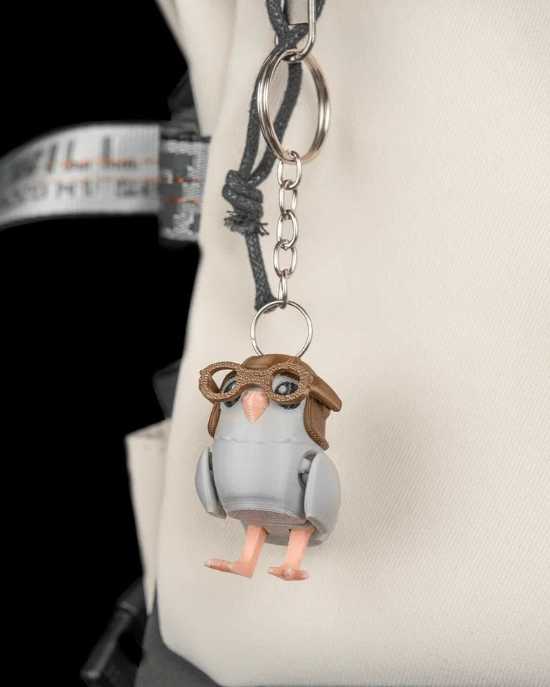 Small gray pigeon figure attached to a metal ring and chain against a light-colored bag