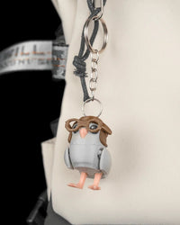Small gray pigeon figure attached to a metal ring and chain against a light-colored bag