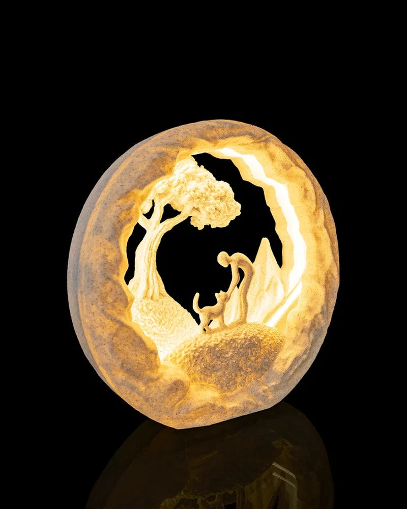 Angled view of a warm glowing round table lamp with a cat, person, tree and mountains