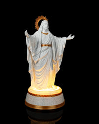 Angled front view of a white and gold Christ statue with outstretched arms and a glowing base