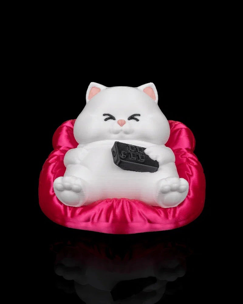 White cat figurine reclining on a bright pink cushion and holding a black remote on a black background.