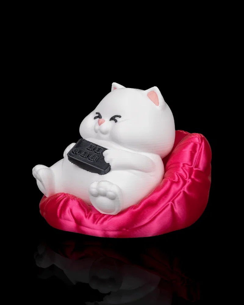 Angled view of a white cat figurine on a bright pink cushion with a black remote in its paws.
