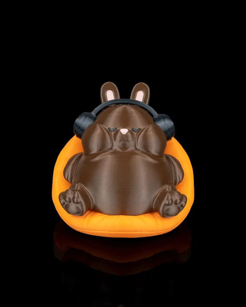 Front view of a brown rabbit figurine wearing black headphones while sitting in an orange carrot-shaped seat on a black background.