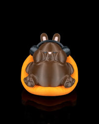 Front view of a brown rabbit figurine wearing black headphones while sitting in an orange carrot-shaped seat on a black background.