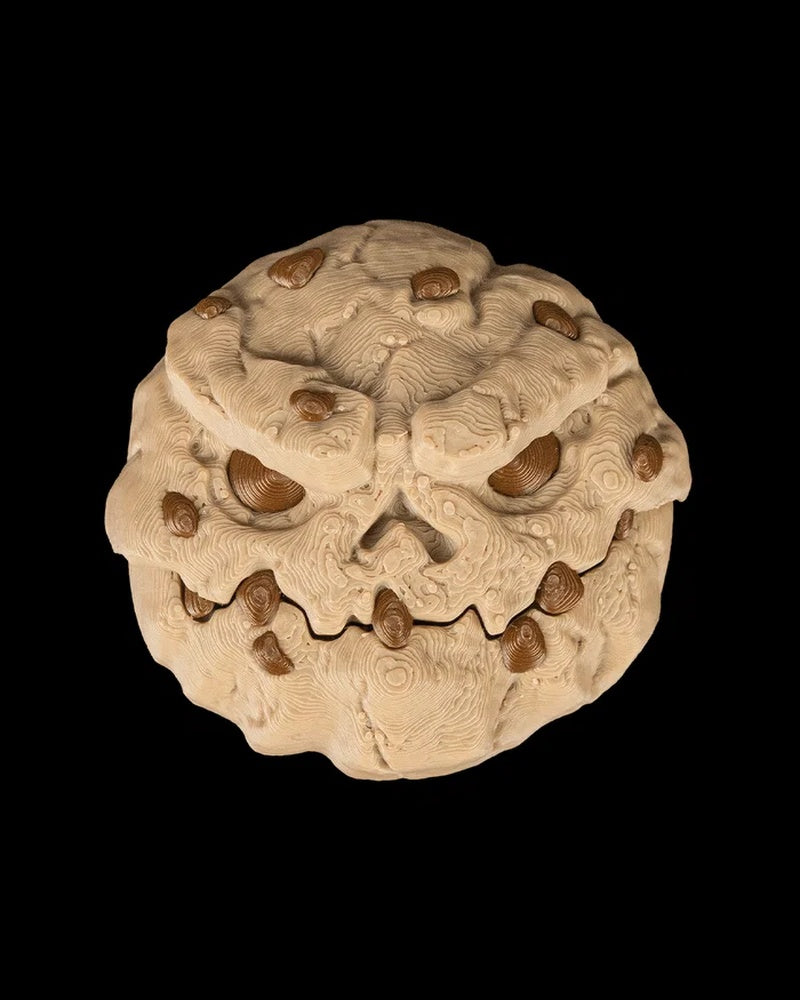 Front view of a tan round monster face with brown chip-like spots, hollow eyes, and a jagged grin on a black background.