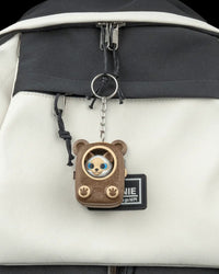 Small brown pack-shaped cat figure hanging by a metal ring and chain from a light-colored bag