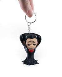 A hand holding a metal ring attached by a short chain to a small black-collared monster figure with a pale face and red accent.