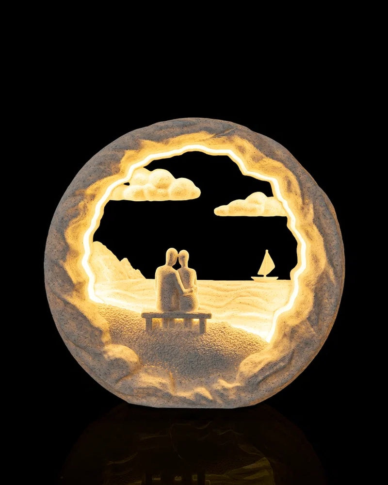 Round illuminated table lamp with two seated figures on a bench, waves, clouds, and a sailboat on a black background