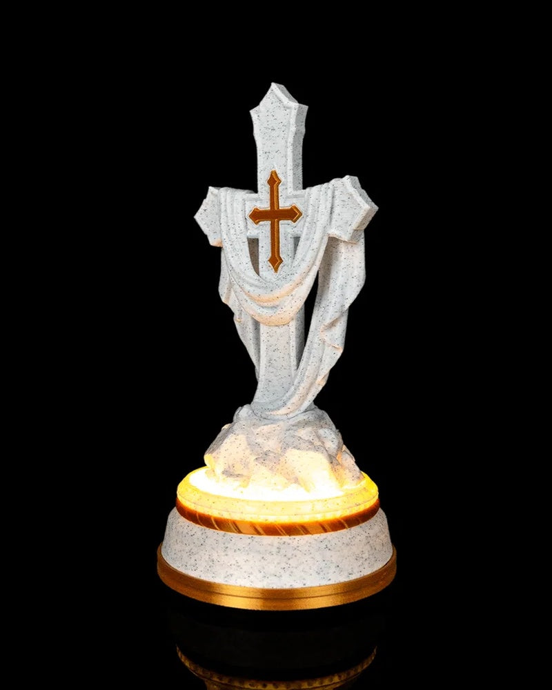 Angled front view of a white speckled draped cross statue with a gold-tone accent and warm glow around the base.