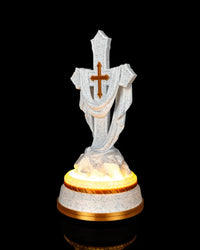 Angled front view of a white speckled draped cross statue with a gold-tone accent and warm glow around the base.