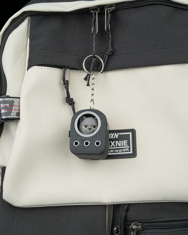 Small grey cat figure in a backpack-shaped frame hanging from a ring and chain on a bag