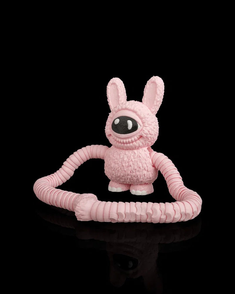 Pink one-eyed monster figurine sitting with long arms looped in front on a black background