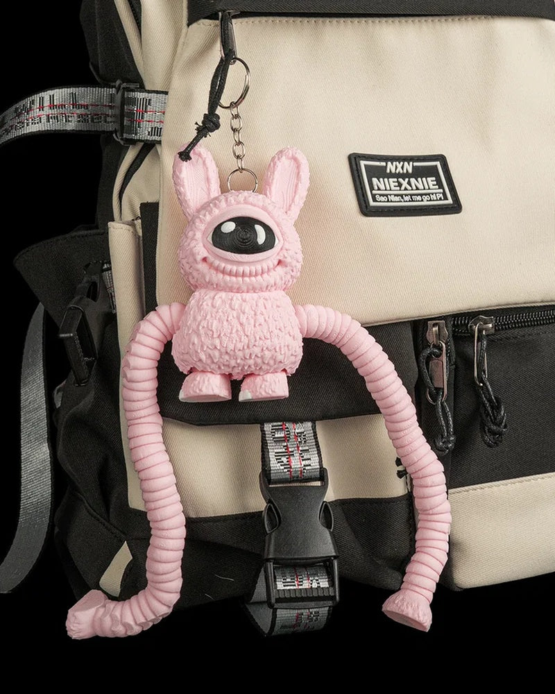 Pink one-eyed monster figure hanging by a metal ring and chain from a beige and black backpack