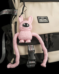 Pink one-eyed monster figure hanging by a metal ring and chain from a beige and black backpack