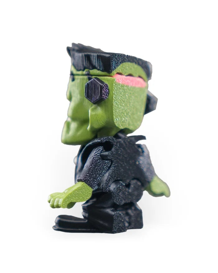 Left side view of a green monster figurine with black flat hair, neck bolt, black outfit, and pink detail at the back of the head.