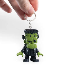 A hand holds a metal key ring attached to a small green monster figurine with black hair and black clothing.