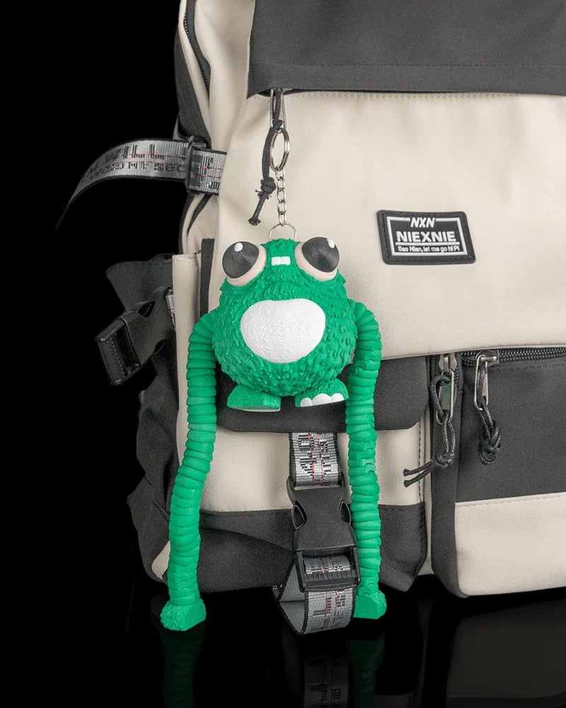Green frog figure hanging from a metal ring and chain on a beige and black backpack