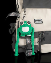 Green frog figure hanging from a metal ring and chain on a beige and black backpack