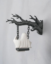 Back view of a white ghost figurine seated on a black swing beneath a dark branch-like support