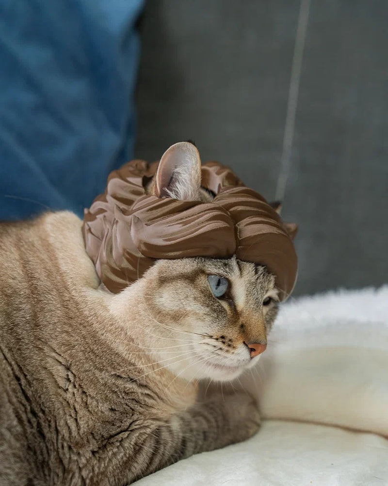 Tabby cat in side view wearing a brown wave-styled headpiece on a white blanket.