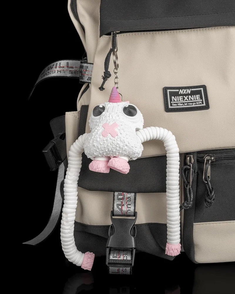 White and pink monster figure hanging by a metal ring and chain from a beige and black backpack