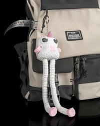 White and pink monster figure with long legs hanging from a beige and black backpack