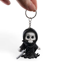 A hand holds a metal ring and short chain above a small black hooded skull figure with a gray blade-like prop.