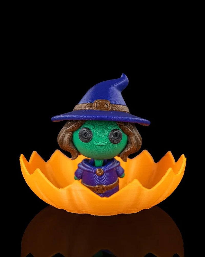 Small witch character in a blue hat sitting inside a jagged orange pumpkin shell on a black background