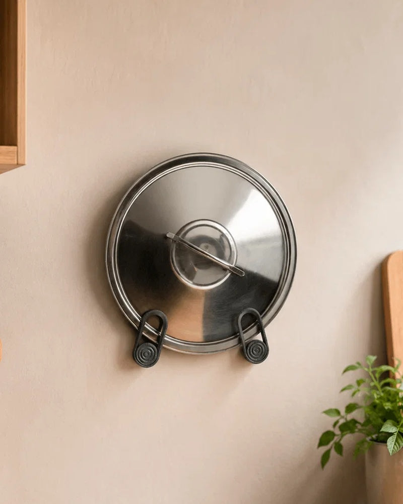 Metal pot lid resting in two black wall-mounted loop holders on a kitchen wall