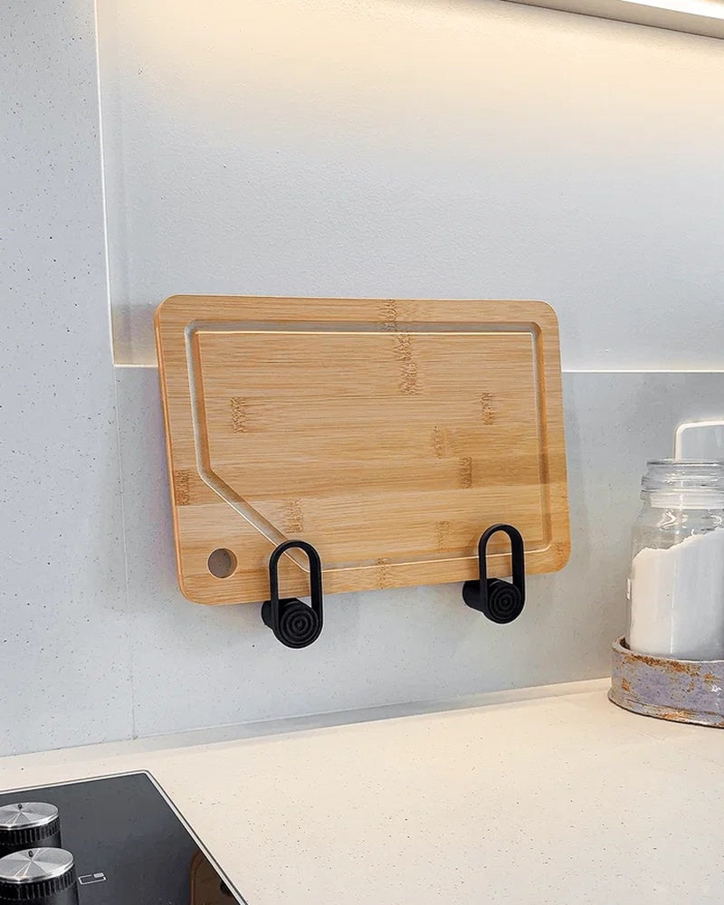 Wood cutting board resting in two black wall-mounted loop holders above a kitchen counter