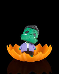Green monster figurine sitting inside an orange jagged pumpkin shell on a black background