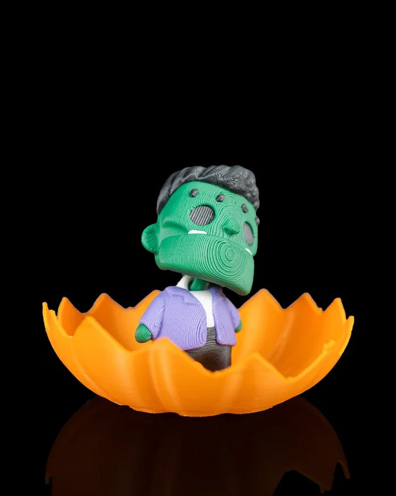 Green monster figurine in a purple jacket inside an orange pumpkin shell on a black background
