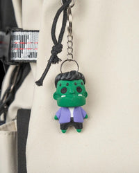 Small green monster figure hanging from a metal ring and chain against a light fabric background