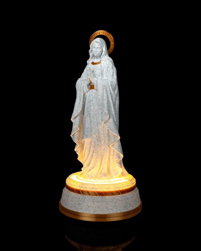 Angled front view of the statue with folded hands, gold halo, and warm light at the base.