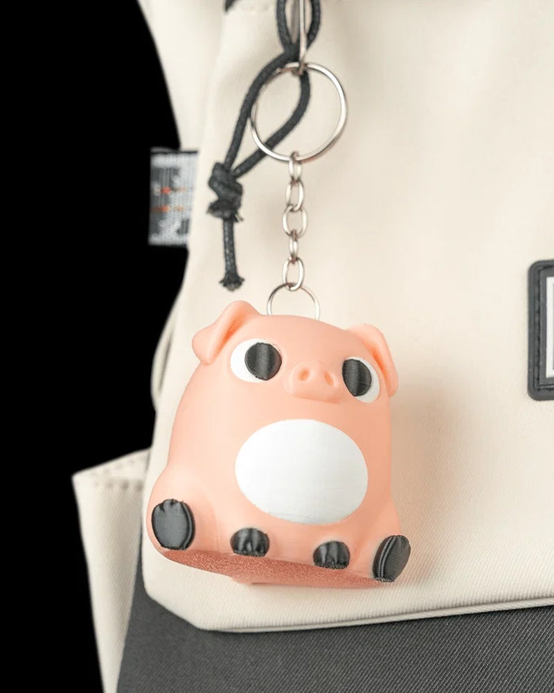 A peach pig keychain hangs from a metal ring on a light-colored bag, showing a white belly, black eyes, and dark hooves.