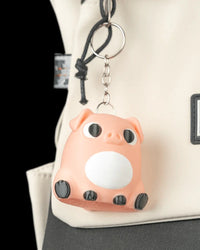 A peach pig keychain hangs from a metal ring on a light-colored bag, showing a white belly, black eyes, and dark hooves.