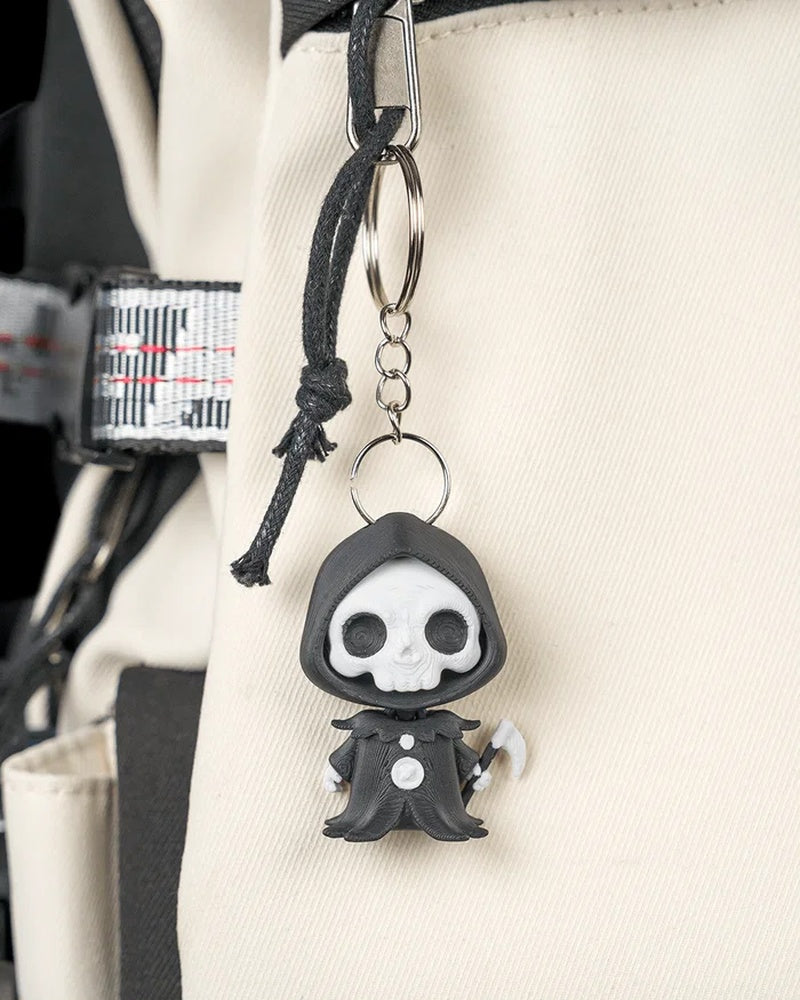 Small hooded skull character hanging from a metal ring and chain on a light-colored bag