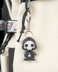 Small hooded skull character hanging from a metal ring and chain on a light-colored bag