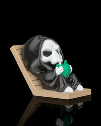Hooded skull character reclining on a tan lounge chair and holding a green drink against a black background