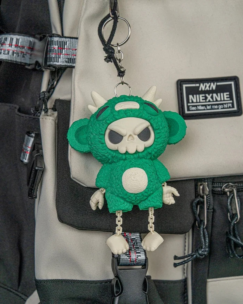 Green skull-faced monster toy with dangling legs hanging from a ring and chain on a bag