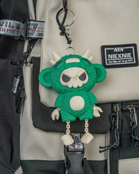 Green skull-faced monster toy with dangling legs hanging from a ring and chain on a bag