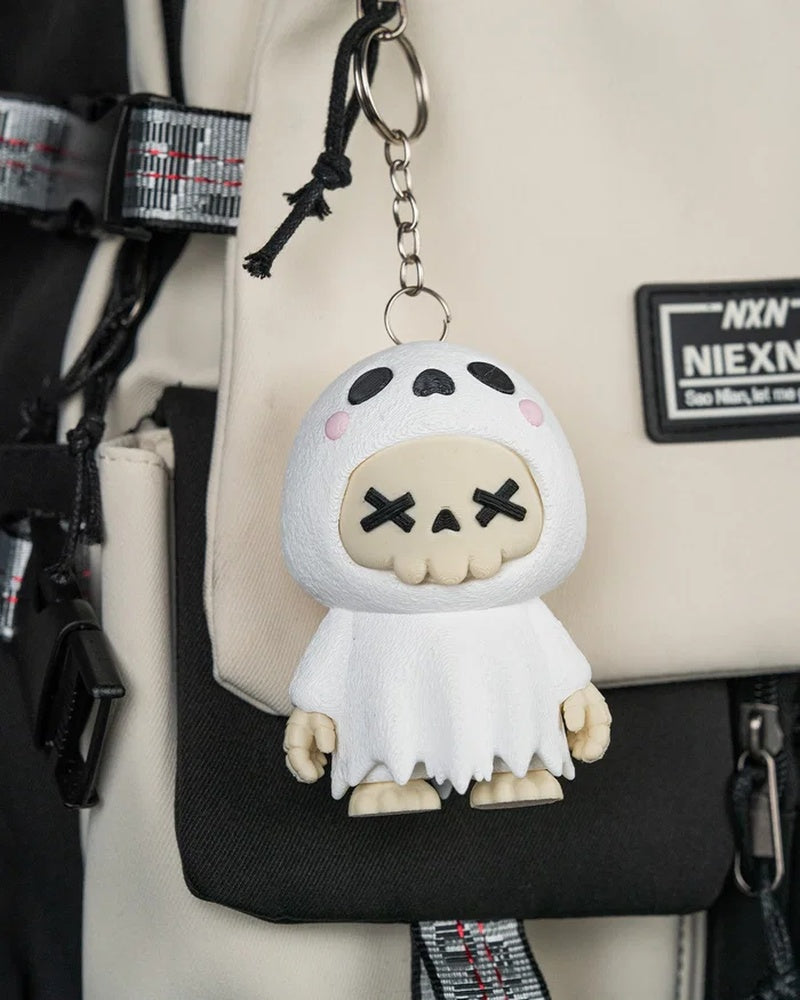 White ghost skull figurine hanging from a metal ring and chain against a beige and black bag.