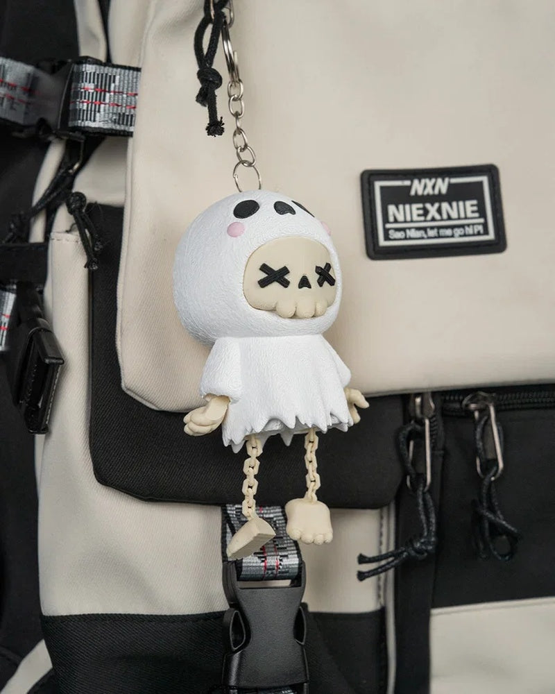 Angled view of the white ghost skull figurine hanging from a metal ring and chain against a beige and black bag.
