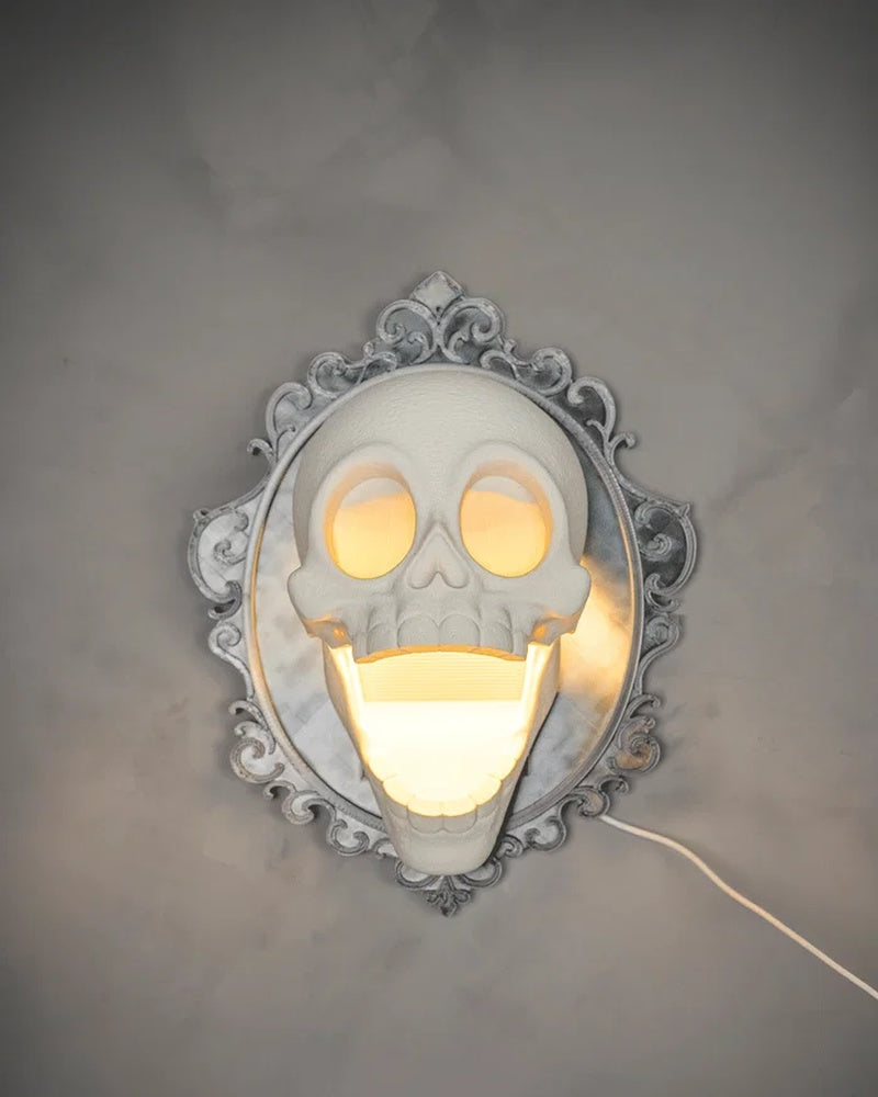 Skull wall lamp glowing on a gray wall with an ornate frame and visible cord