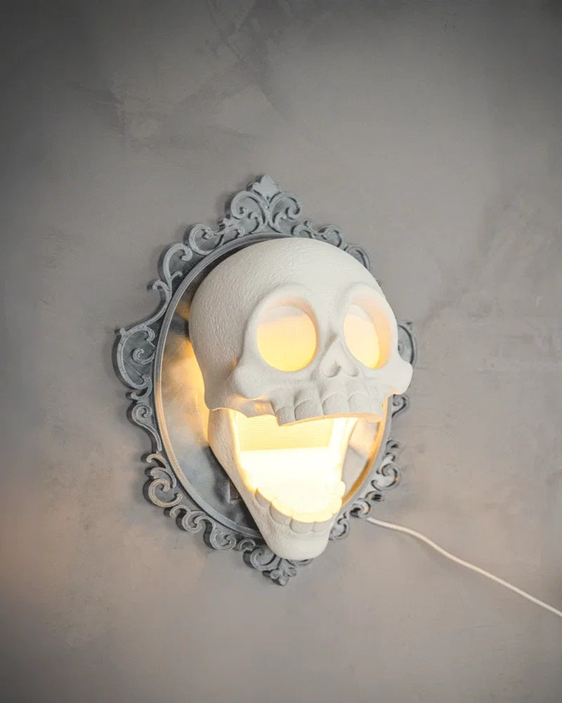 Angled view of a glowing skull wall lamp on a gray wall with an ornate frame