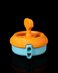 Orange and blue circular toy with a raised top grip and front whistle-shaped opening on a black background