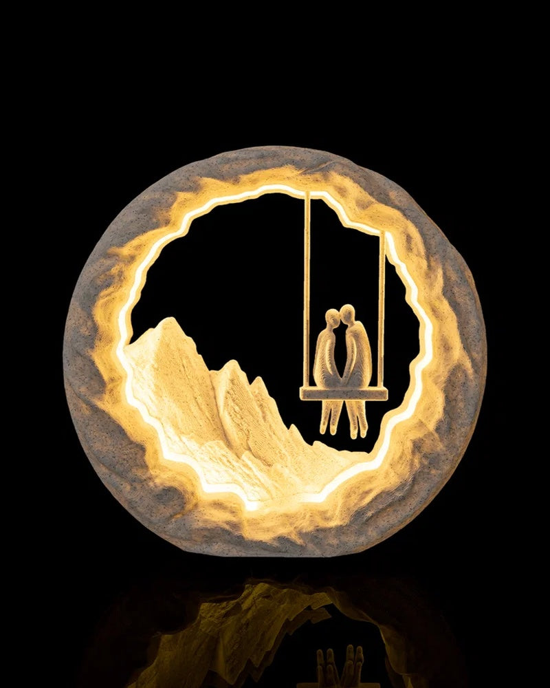 Round glowing table lamp with two figures seated on a hanging swing and mountain-like forms inside