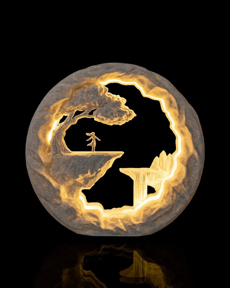Round illuminated table lamp with a cliffside scene, small figure, tree and warm glowing cutouts on a dark background