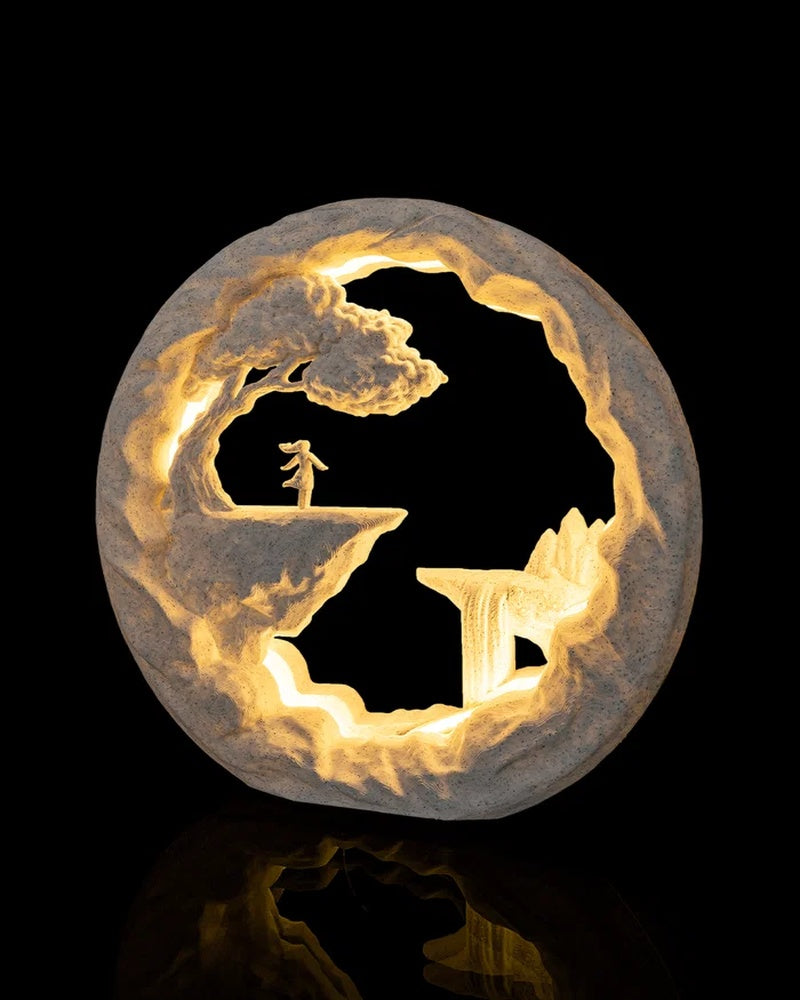 Round illuminated landscape lamp with a small figure under a tree and warm light around rocky cutouts