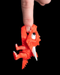 Red dragon toy held upright by a finger with its mouth open against a black background